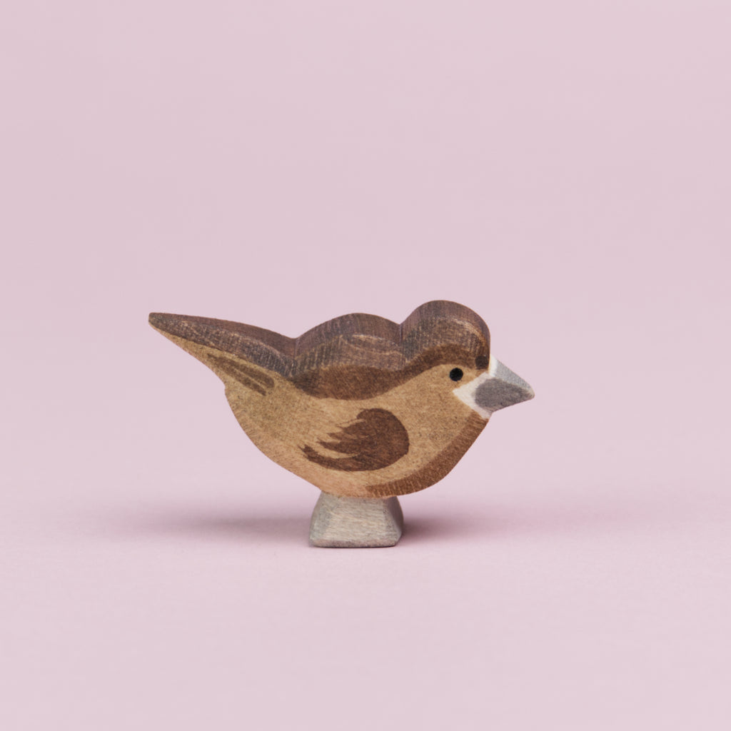 Ein Spatz aus Holz in von der Seite zu sehen und schaut nach rechts. Seine Federn sind hellbraun, auf dem Kopf und dem Rücken bis hin zu den Schwanzfedern und auch an den Flügeln hat er dunkelbraune Federn. Der Schnabel und die Füße sind grau bemalt und um den Schnabel herum ist eine weiße Linie zu sehen. Der Spatz ist von der Marke Ostheimer.