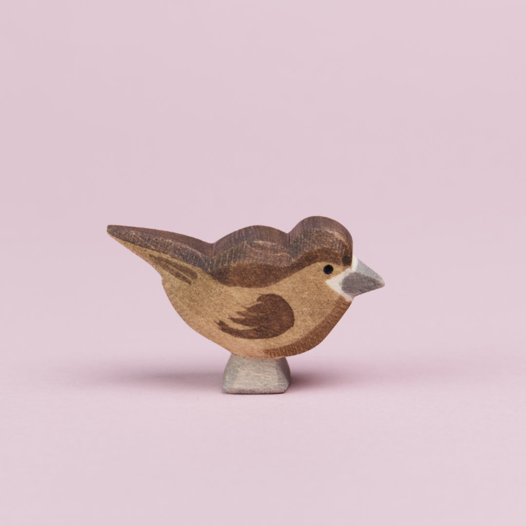 Ein Spatz aus Holz in von der Seite zu sehen und schaut nach rechts. Seine Federn sind hellbraun, auf dem Kopf und dem Rücken bis hin zu den Schwanzfedern und auch an den Flügeln hat er dunkelbraune Federn. Der Schnabel und die Füße sind grau bemalt und um den Schnabel herum ist eine weiße Linie zu sehen. Der Spatz ist von der Marke Ostheimer.