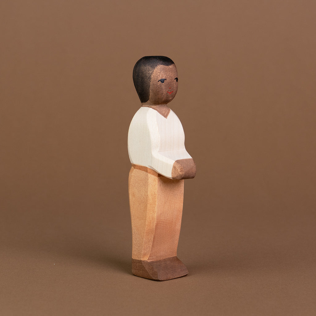 Ein Vater aus Holz von der Marke Ostheimer mit dunkler Hautfarbe, dunkelbraunen Haaren und Augen ist leicht eingedreht, stehend nach rechts vorne schauend, zu sehen. Seine beiden Arme hält er nach vorne zusammen und er trägt ein helles Oberteil mit V-Ausschnitt, eine hellbraune Hose mit einem braunen Gürtel und dunkelbraune Schuhe.