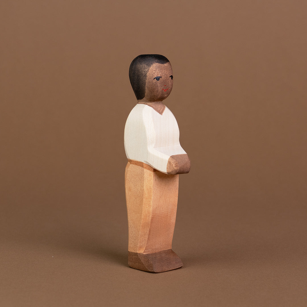 Ein Vater aus Holz von der Marke Ostheimer mit dunkler Hautfarbe, dunkelbraunen Haaren und Augen ist leicht eingedreht, stehend nach rechts vorne schauend, zu sehen. Seine beiden Arme hält er nach vorne zusammen und er trägt ein helles Oberteil mit V-Ausschnitt, eine hellbraune Hose mit einem braunen Gürtel und dunkelbraune Schuhe.