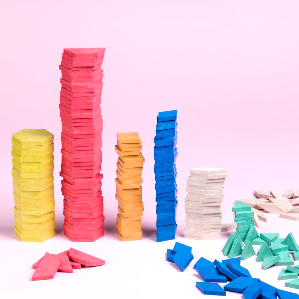 Die Pattern blocks Teil, im Set 250 Stück enthalten, sind teils farblich in gelb, rot, orange, blau, natur und grün, in unterschiedlichen Formen, 6 Eck, Trapez, Quadrat, Raute und Dreieck, in unterschiedlichen Größen aufeinander gestapelt. Ein paar Steine liegen lose daneben.
