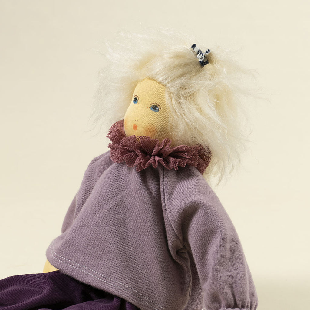 Nanchen Puppe mit heller Haut und hellblonde Haaren sitzend. Sie trägt einen Pullover in Flieder mit Kragen und eine lila Hose.