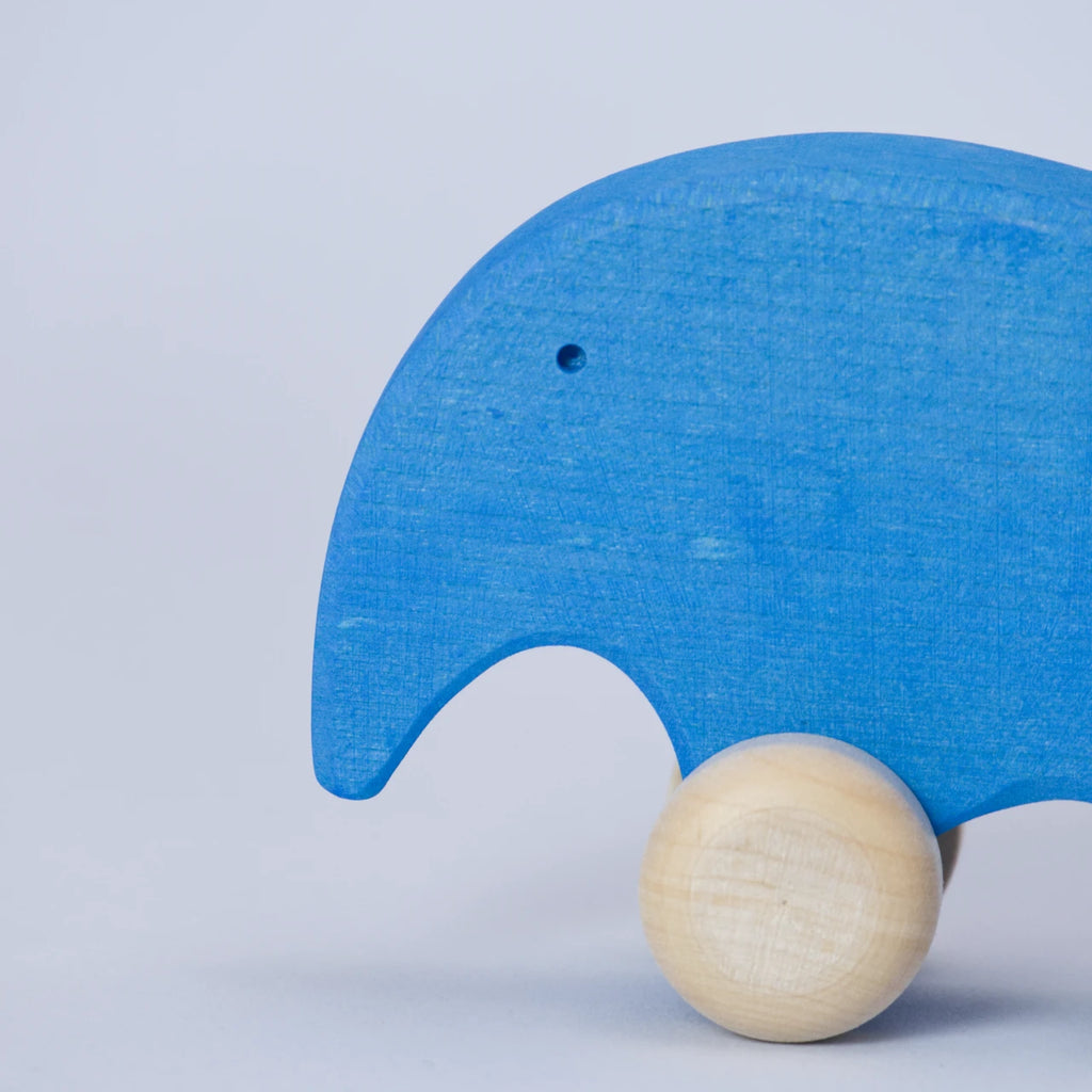 Ein blaues Schiebetier aus Holz mit weißen Rädern von Goki Evolution ist in einer Nahaufnahme zu sehen. Die Form erinnert abstrahiert an einen Elefanten, dieser schaut nach links. Sein Auge ist ein kleines Loch.