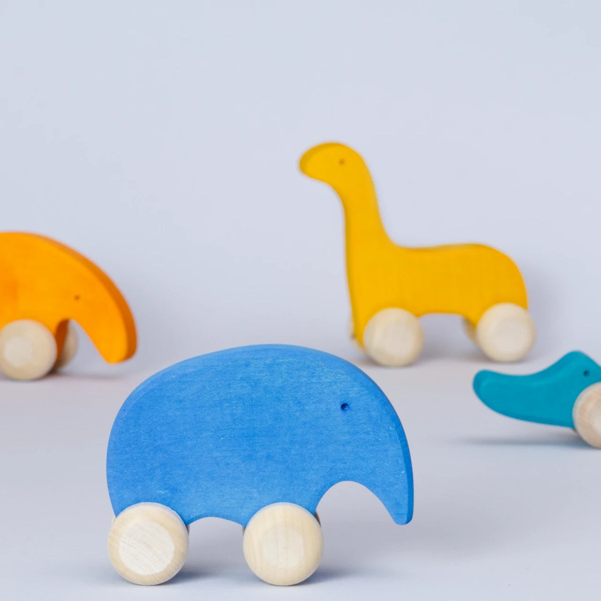 Ein Gruppenbild von den Holz Schiebetieren von Goki Evolution. Der blaue Elefant steht im Vordergrund und schaut nach rechts. Der orangene Ameisenbär, der gelbe Dinosaurier und das grüne Krokodil sind unscharf im Hintergrund.