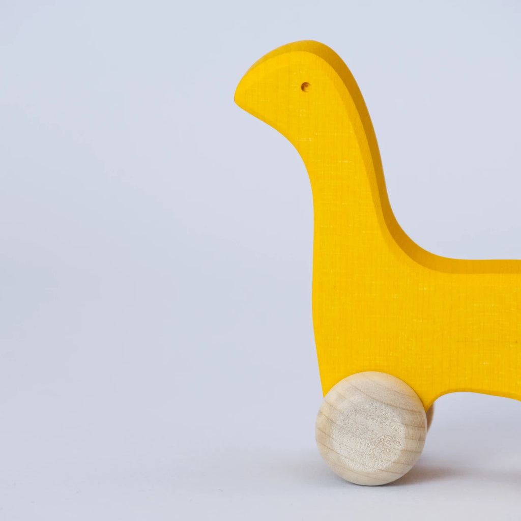 Ein gelbes Schiebetier mit weißen Rädern von Goki Evolution ist in einer Nahaufnahme im Profil zu sehen. Die Form erinnert an einen Dinosaurier mit langem Hals oder einer Giraffe. Das Tier schaut nach links. Es ist aus Holz gefertigt und die Farbe ist ein intensives, freundliches Gelb.