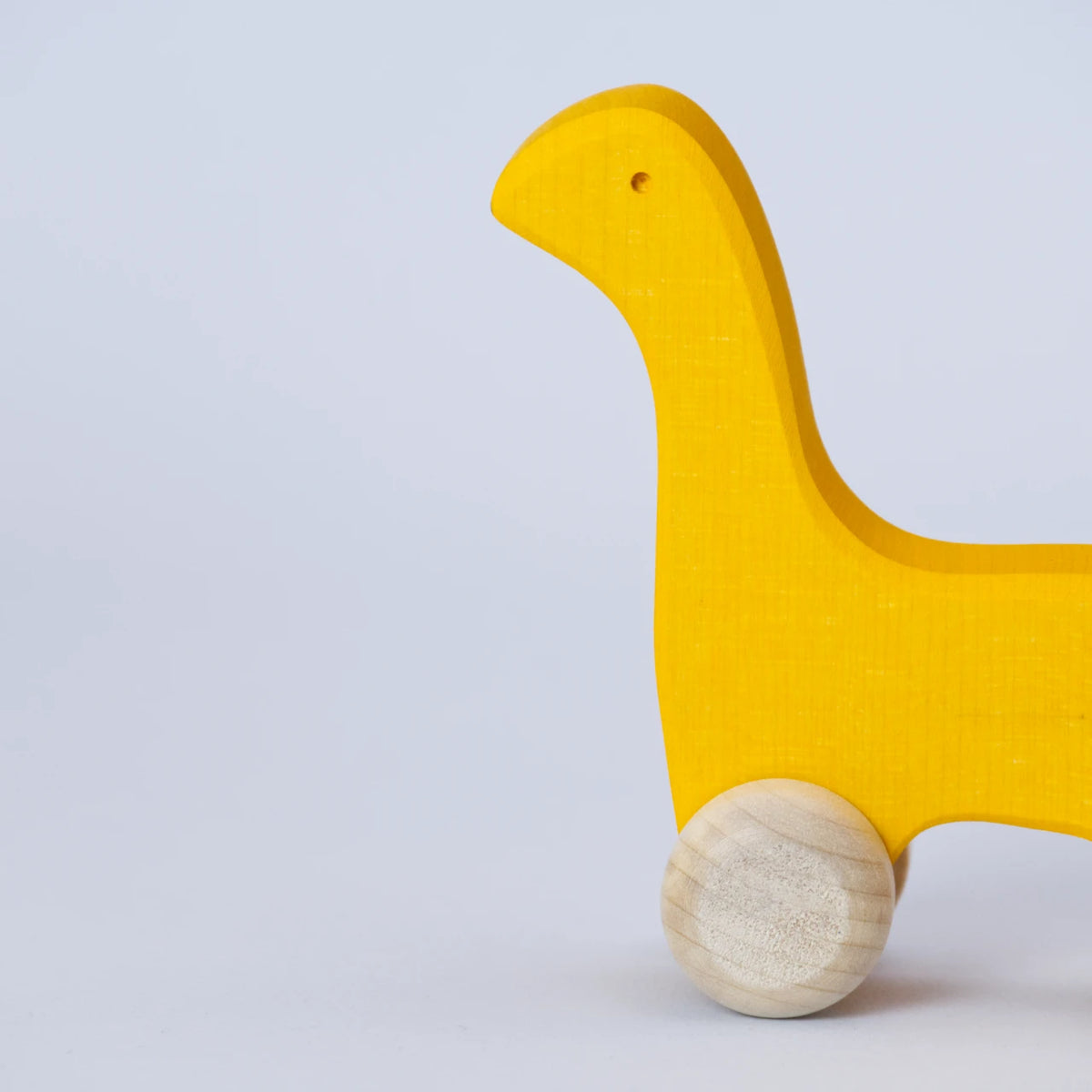 Ein gelbes Schiebetier mit weißen Rädern von Goki Evolution ist in einer Nahaufnahme im Profil zu sehen. Die Form erinnert an einen Dinosaurier mit langem Hals oder einer Giraffe. Das Tier schaut nach links. Es ist aus Holz gefertigt und die Farbe ist ein intensives, freundliches Gelb.