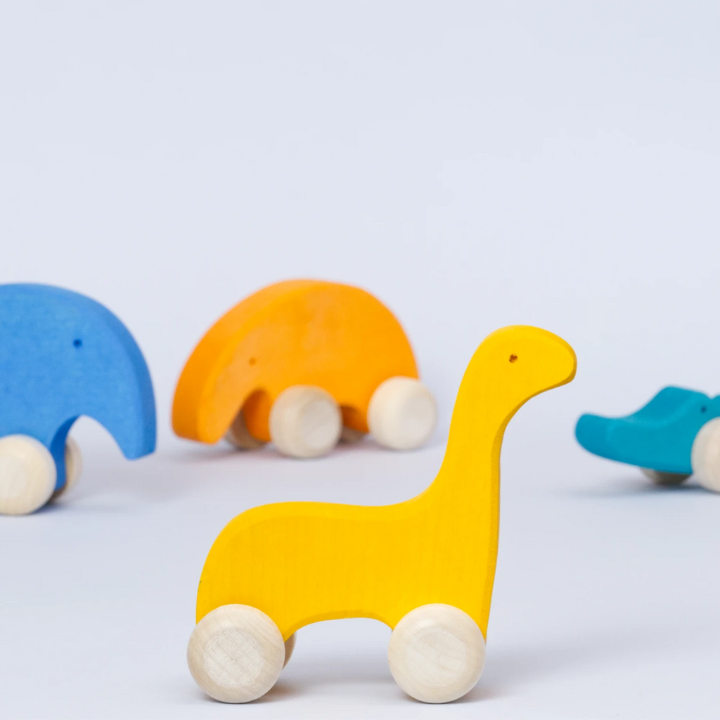 Ein Gruppenbild von den Holz Schiebetieren von Goki Evolution. Der gelbe Dinosaurier steht im Vordergrund und schaut nach rechts.Der blaue Elefant, der orangene Ameisenbär und das grüne Krokodil sind unscharf im Hintergrund.