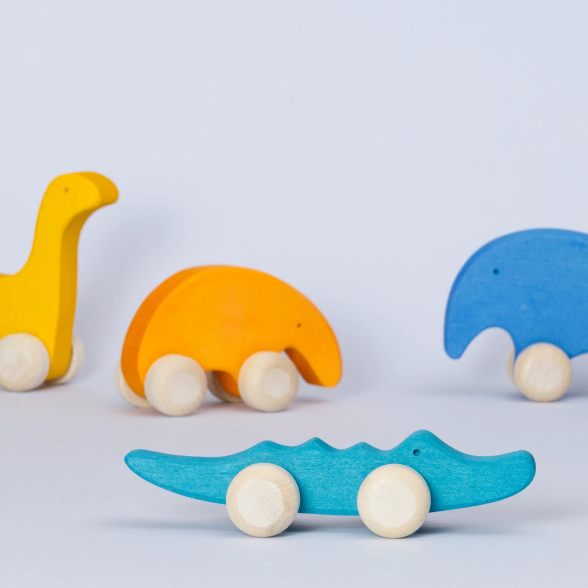 Ein Gruppenbild von den Holz Schiebetieren von Goki Evolution. Das grüne Krokodil steht im Vordergrund und schaut nach rechts. Der gelbe Dinosaurier, der orangene Ameisenbär und der blaue Elefant sind unscharf im Hintergrund.