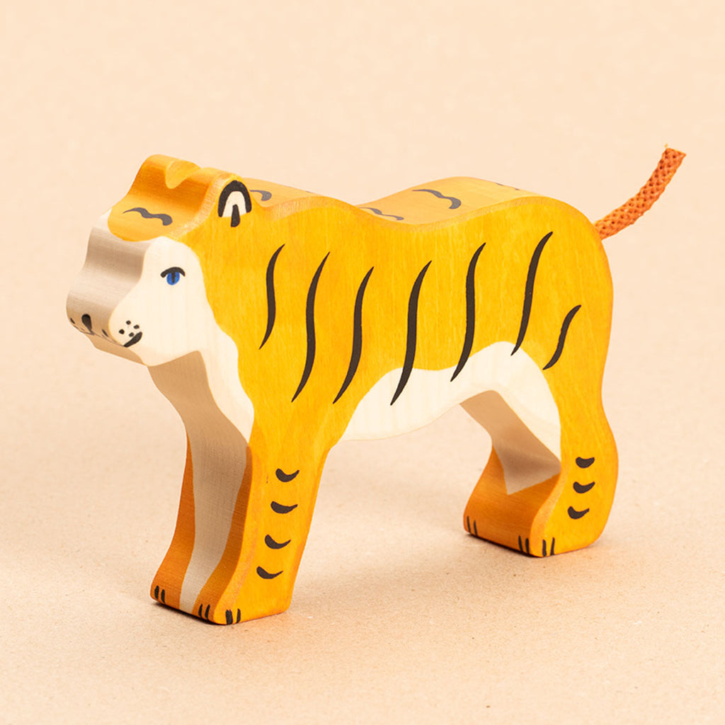 Ein nach vorne links schauender Tiger aus Naturholz, hat orangenes Fell mit senkrechten, schwarzen Streifen. Sein Gesicht und Bauchfell sind weiß. Sein orangener Schwanz aus Kordel zeigt nach oben hin. Sein Auge ist blau aufgemalt. Die schwarzen Streifen an den Beinen sind jedoch waagerecht aufgemalt.
