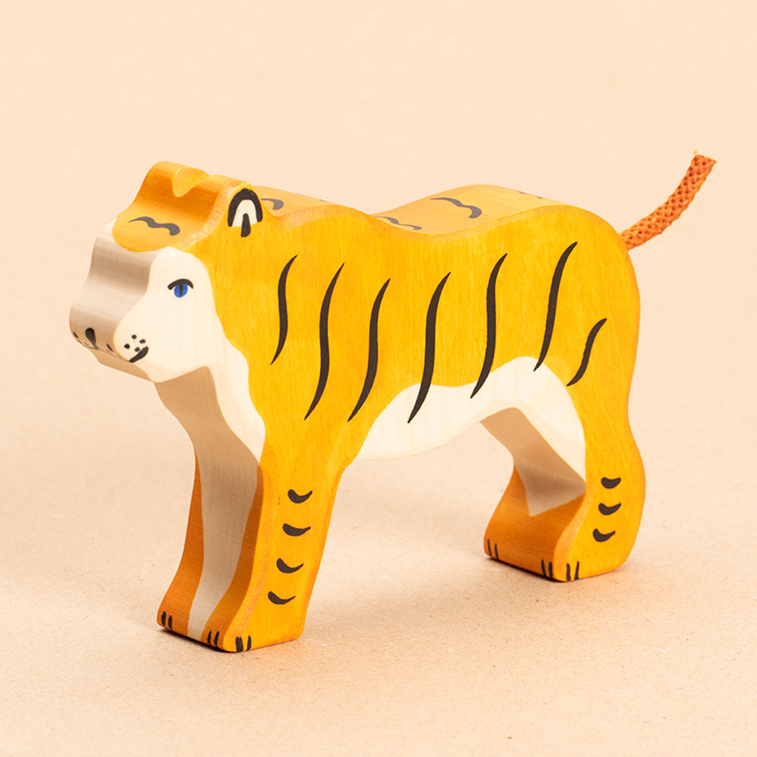 Ein nach vorne links schauender Tiger aus Naturholz, hat orangenes Fell mit senkrechten, schwarzen Streifen. Sein Gesicht und Bauchfell sind weiß. Sein orangener Schwanz aus Kordel zeigt nach oben hin. Sein Auge ist blau aufgemalt. Die schwarzen Streifen an den Beinen sind jedoch waagerecht aufgemalt.