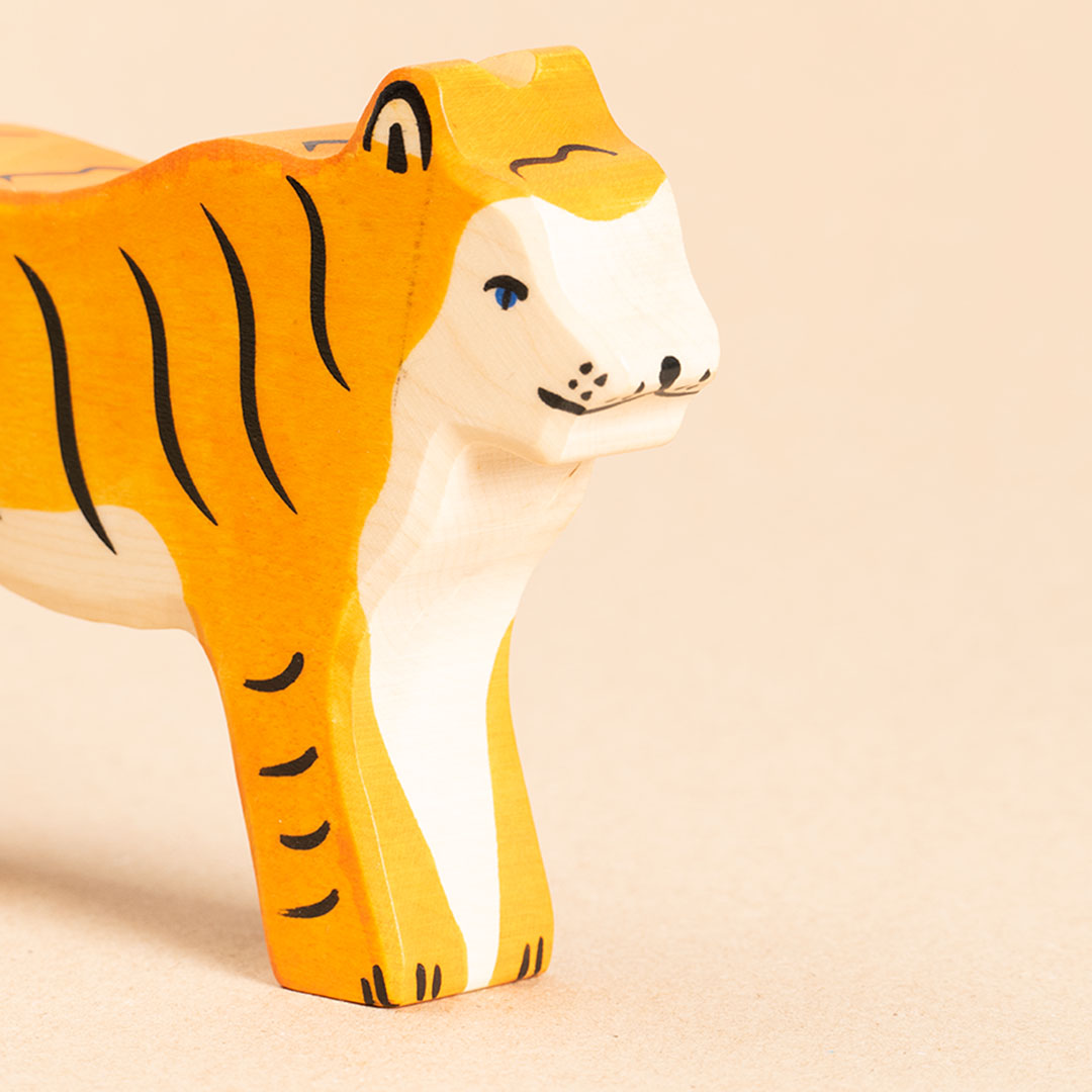 Die Nahaufnahme zeigt einen orangenen, nach vorne rechts schauenden Holztiger mit blauen Augen. Das Fell ist mit schwarzen, senkrechten Streifen versehen. Die Beine weisen hingegen waagerechte, aufgemalte Streifen auf. Sein Bauchfell ist weiß. Sichtbar ist sein Körper bis zur Körpermitte hin.