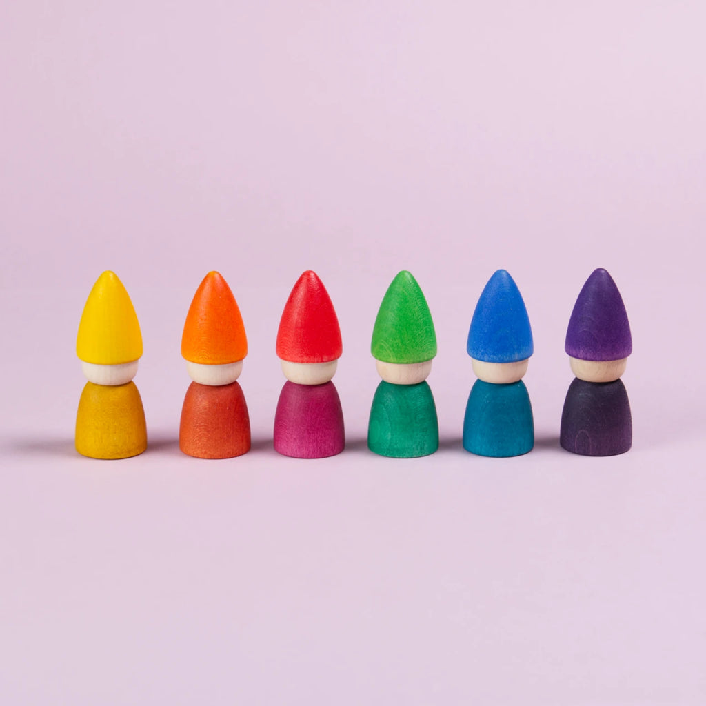 Mittig im Bild nebeneinander aufgereihte Figuren mit einer spitzen Mütze und mit farblich gleichem Oberkörper. In gelb, orange, rot, grün, blau und lila. Alle haben kein Gesicht.