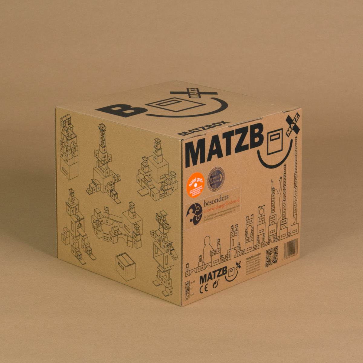 Die MATZBOX steht in der originalen Verpackung aus Pappe, die noch komplett entfaltet ist. Sichtbar sind drei Seiten: der Deckel und zwei Seiten. Auf der Box sind mögliche Bauanleitungen grafisch als Outlines dargestellt. Außerdem ist das Logo der Box zu sehen. Die Pappe ist braun, der Aufdruck schwarz.