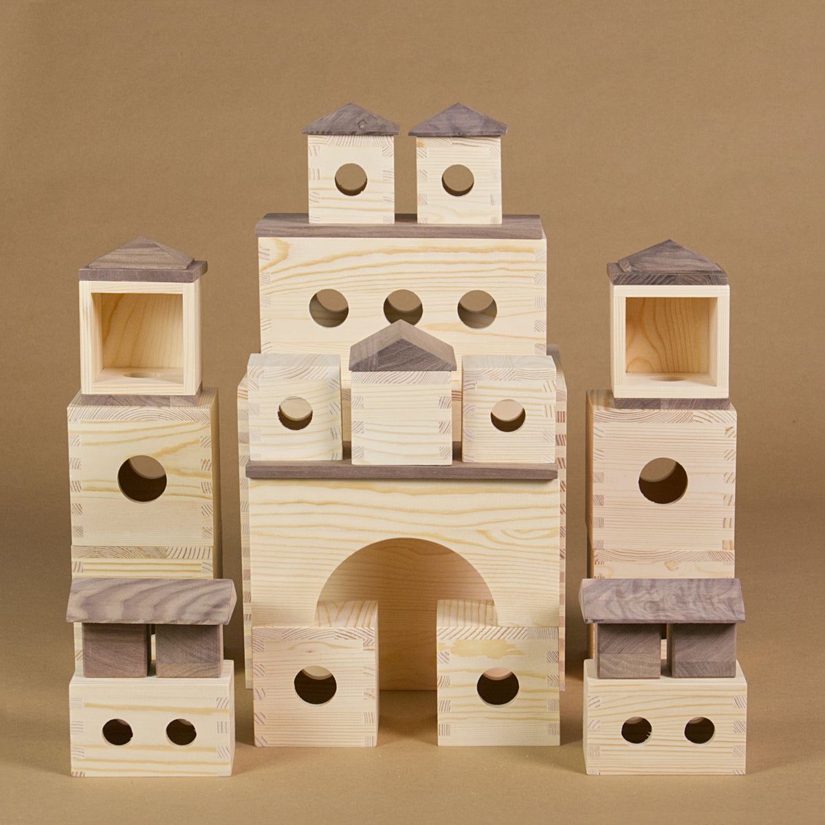 Die MATZBOX ist auf diesem Bild zu einem symmetrischen Gebäude aufgebaut, wobei alle 36 Holzelemente sichtbar sind. Zu erkennen sind offene Würfel mit kreisrunden Löchern, längliche Bauteile mit drei Löchern und einer offenen Seite sowie rechteckige und quadratische Platten aus dunklem Holz. Vier Holzwürfel und pyramiden- sowie pultdachförmige Bausteine ergänzen die Vielfalt. Zwei Torbögen aus hellem Massivholz sind ebenfalls zu sehen.