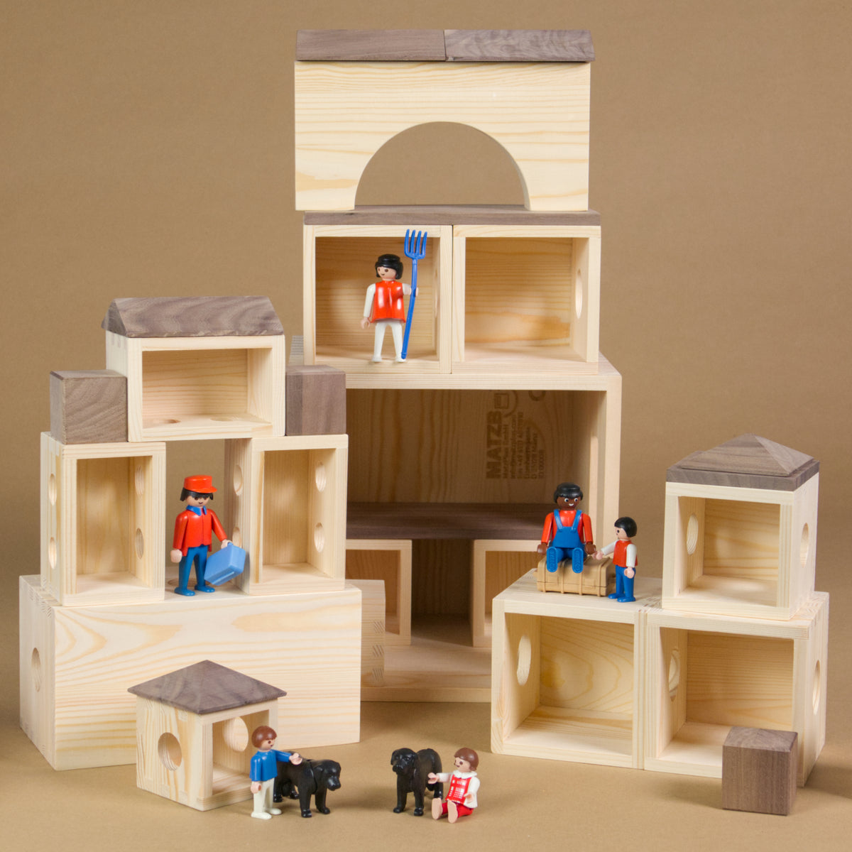 MATZBOX-Idee: Die Holzelemente der MATZBOX, offene Holzkästchen und Bausteine, sind zu einer Sammlung kleinerer Gebilde arrangiert. Die unterschiedlich großen Spielhäuser stehen verteilt. Als Spielfiguren wurden Playmobil-Figuren verwendet. Drei erwachsene Figuren, drei Kinder und zwei Hunde beleben die Spielszene.