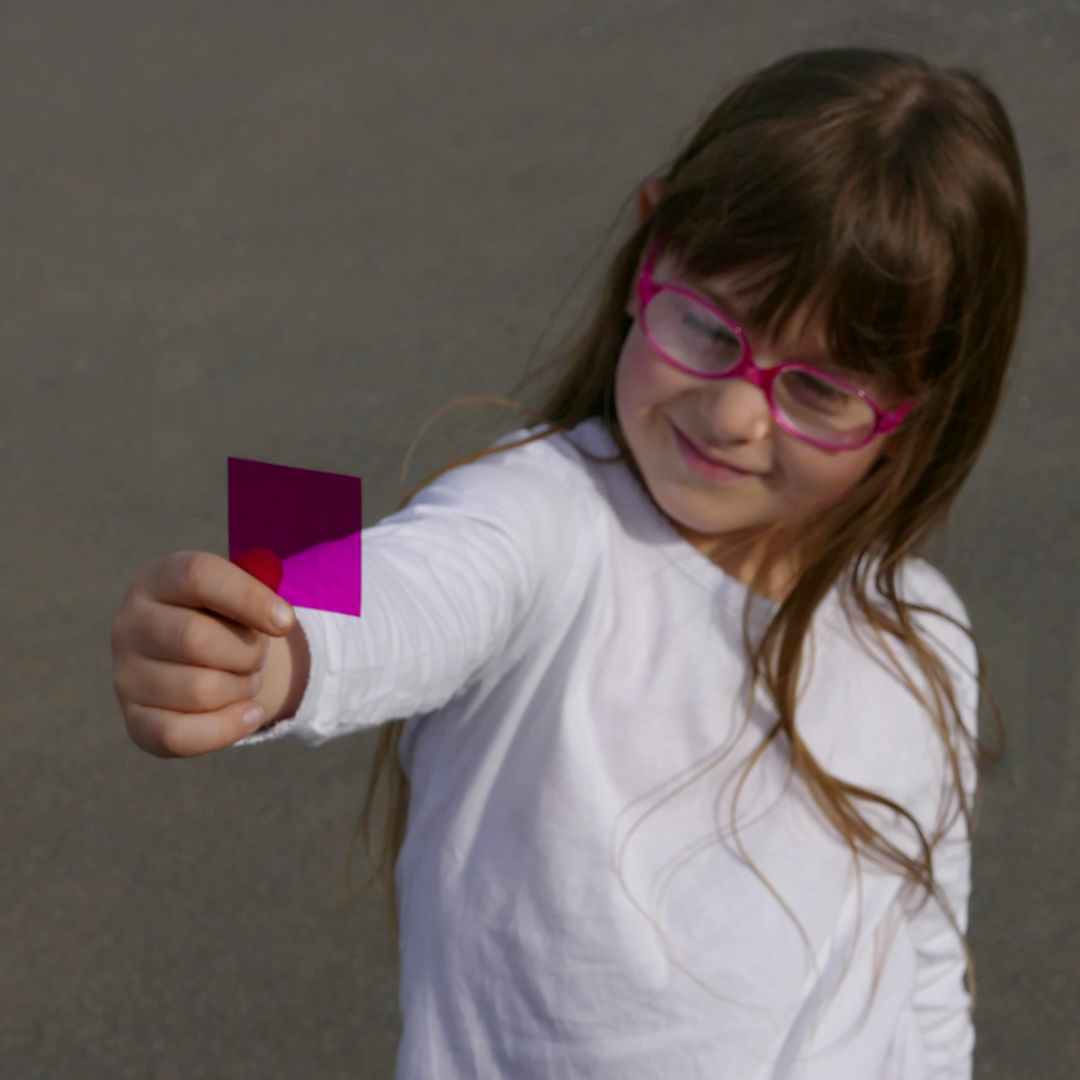 Kind hält quadratisches Farbkärtchen in die Kamera, die pinke Farbkarte ist leicht durchsichtig.