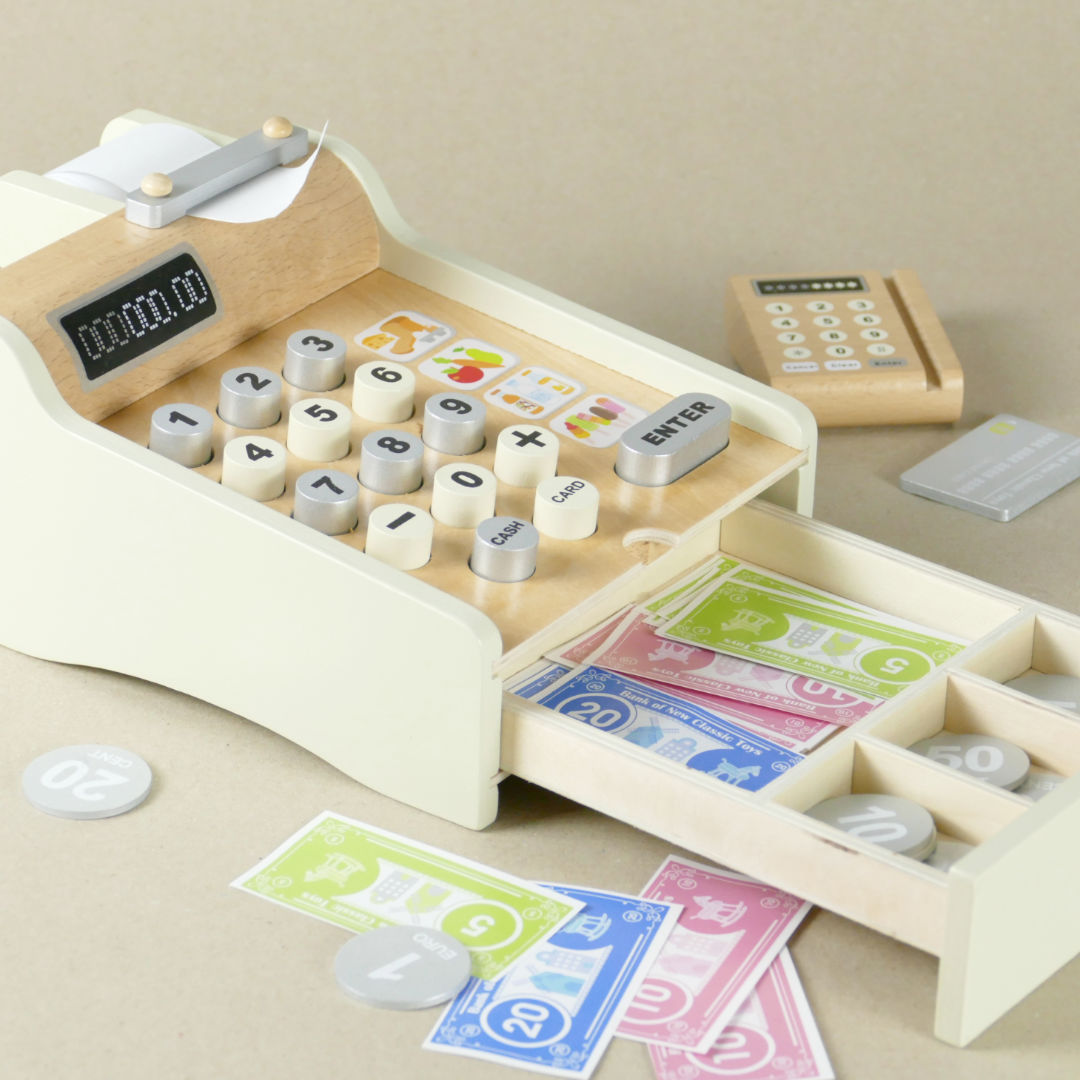 Creme farbene Holz, Spielkasse mit geöffneter Schublade und Spielgeld im Vordergrund. Im Hintergrund Spiellesegerät für Spiel-EC-Karte.