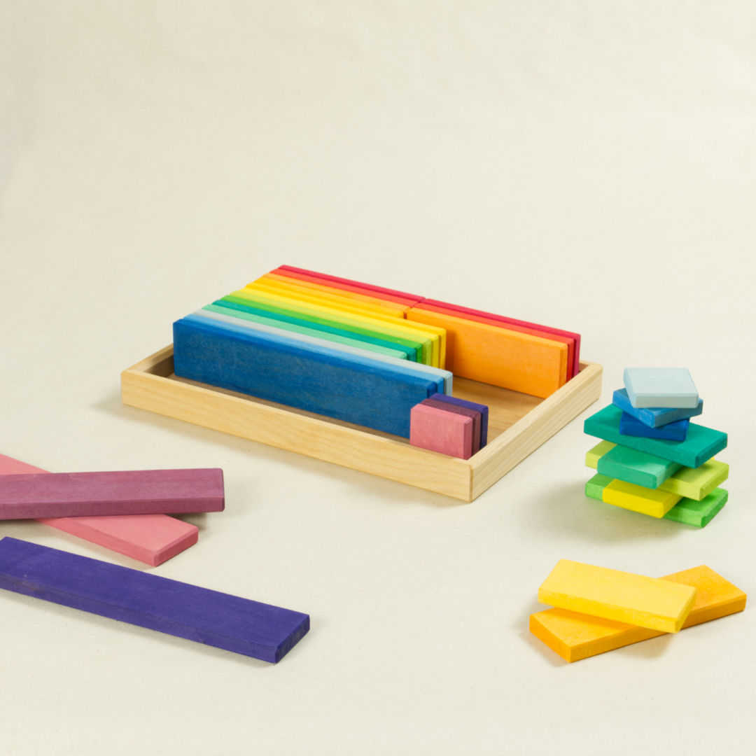 Regenbogenfarbene Bauklötze in unterschiedlichen Längen. Teilweise in Kiste gereiht, auf dem Boden liegend und gestapelt.