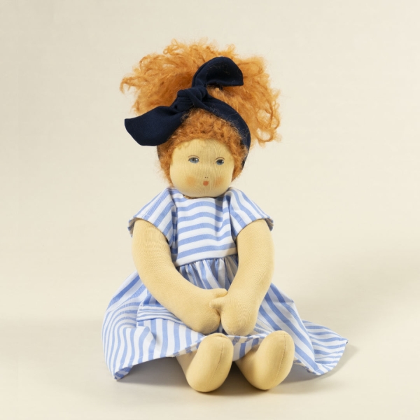Nanchen Puppe mit heller Haut und roten Haaren sitzend. Sie trägt eine blaue Haarschleife und ein blau-weiß gestreiftes Kleid.