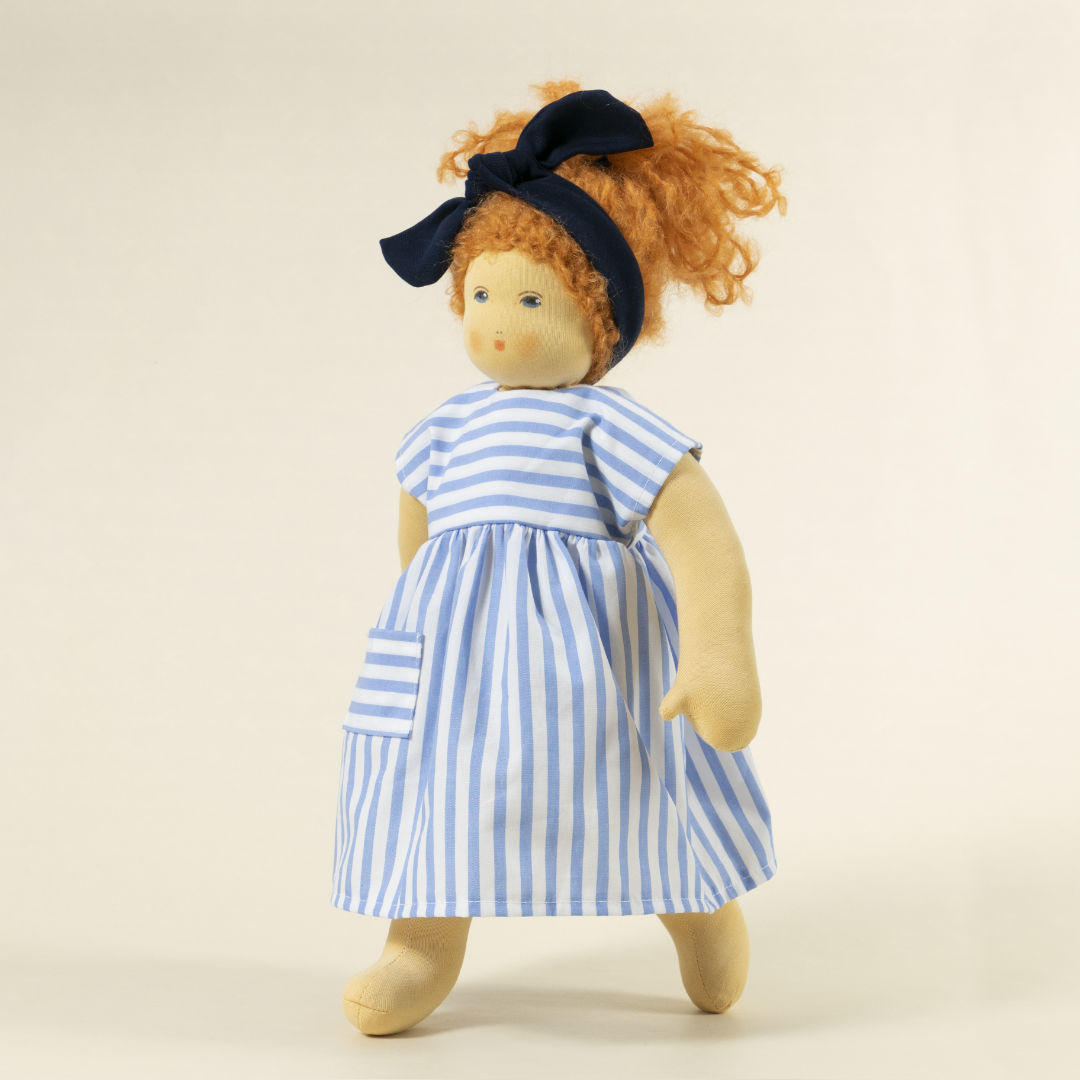 Nanchen Puppe mit heller Haut und roten Haaren stehend. Sie trägt eine blaue Haarschleife und ein blau-weiß gestreiftes Kleid.
