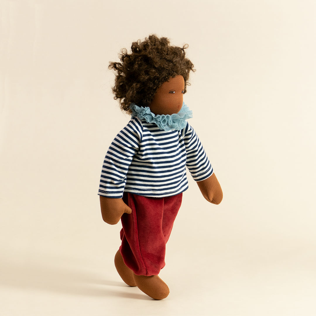Nanchen Puppe mit dunkler Haut und dunkeln Haaren stehend. Sie trägt ein dunkelblau-weiß gestreiftes Oberteil mit Kragen und eine rote Hose.