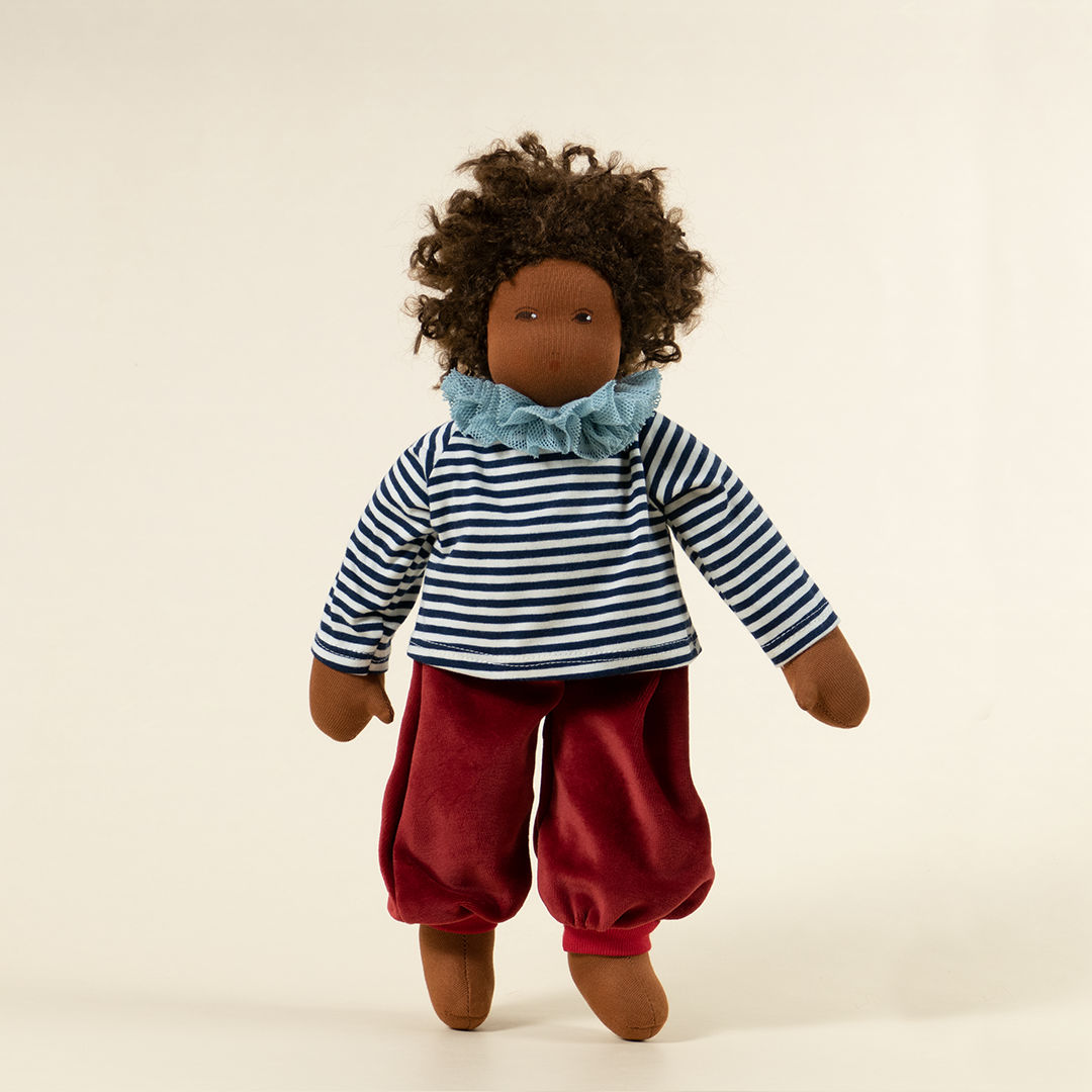 Nanchen Puppe mit dunkler Haut und dunkeln Haaren stehend. Sie trägt ein dunkelblau-weiß gestreiftes Oberteil mit Kragen und eine rote Hose.