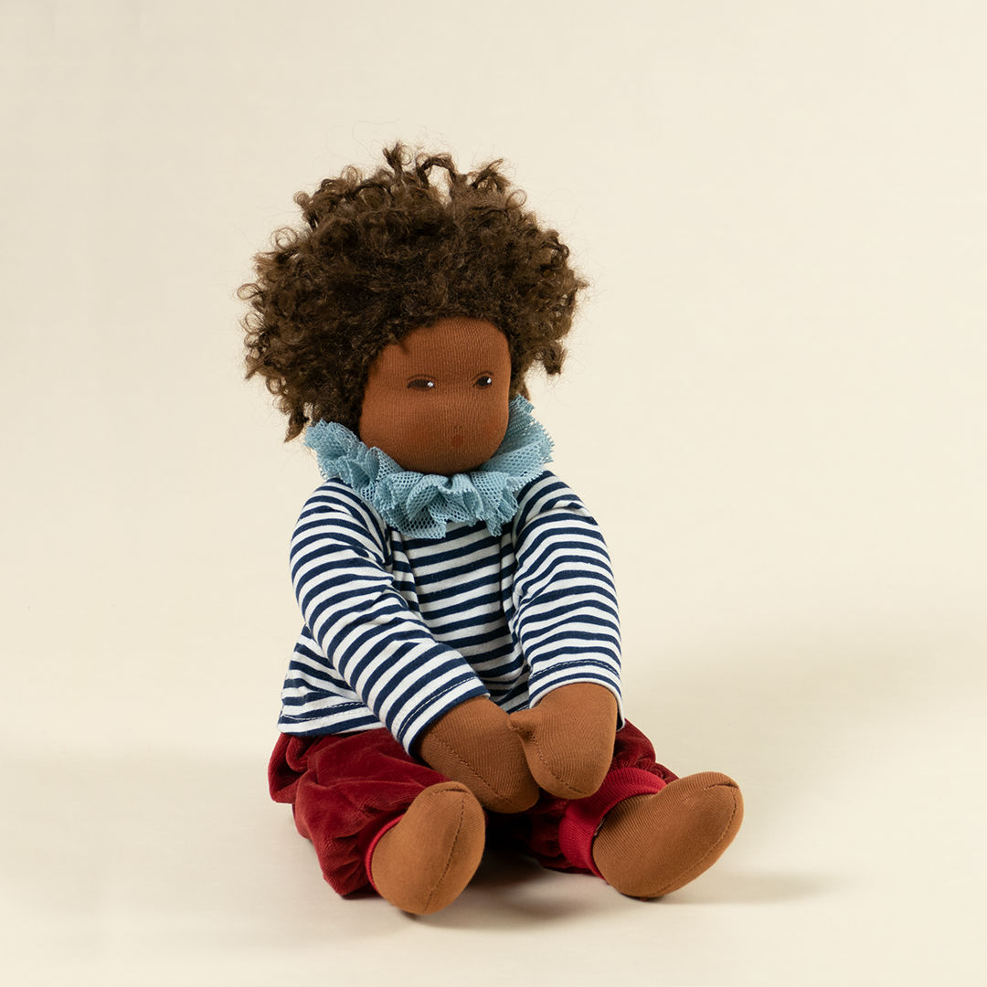 Nanchen Puppe mit dunkler Haut und dunkeln Haaren sitzend. Sie trägt ein dunkelblau-weiß gestreiftes Oberteil mit Kragen und eine rote Hose.