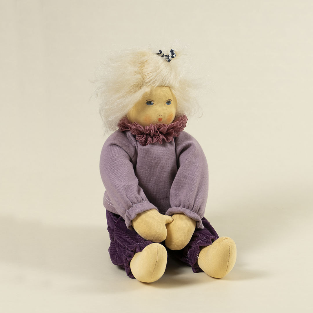 Nanchen Puppe mit heller Haut und hellblonde Haaren sitzend. Sie trägt einen Pullover in Flieder mit Kragen und eine lila Hose.