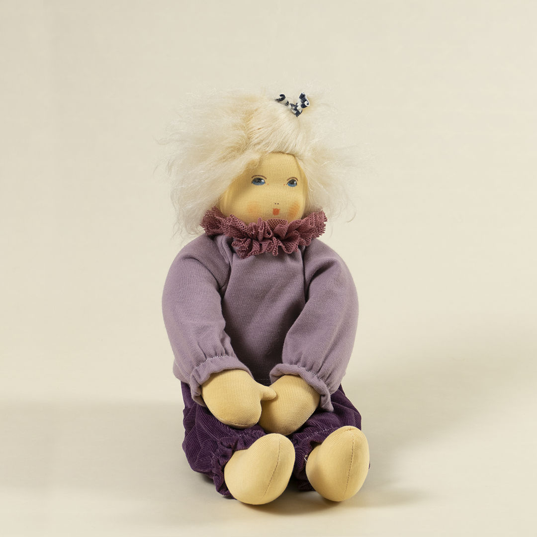 Nanchen Puppe mit heller Haut und hellblonde Haaren sitzend. Sie trägt einen Pullover in Flieder mit Kragen und eine lila Hose.