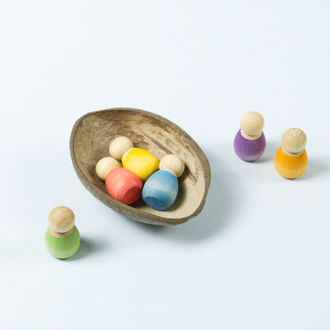 sechs kleine Babyholzfiguren in verschiedenen Farben in einer halben Kokosnussschale liegend und daneben stehend.