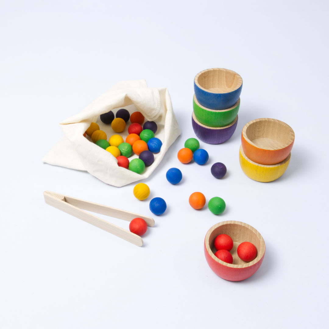 Säckchen mit verschiedenen kleinen Holzkugeln in 6 verschiedenen Farben, daneben gestapelte Holzbecher in auch je 6 Farben und eine Zange um die kleinen Kugeln in die Becher nach Farbe zu sortieren