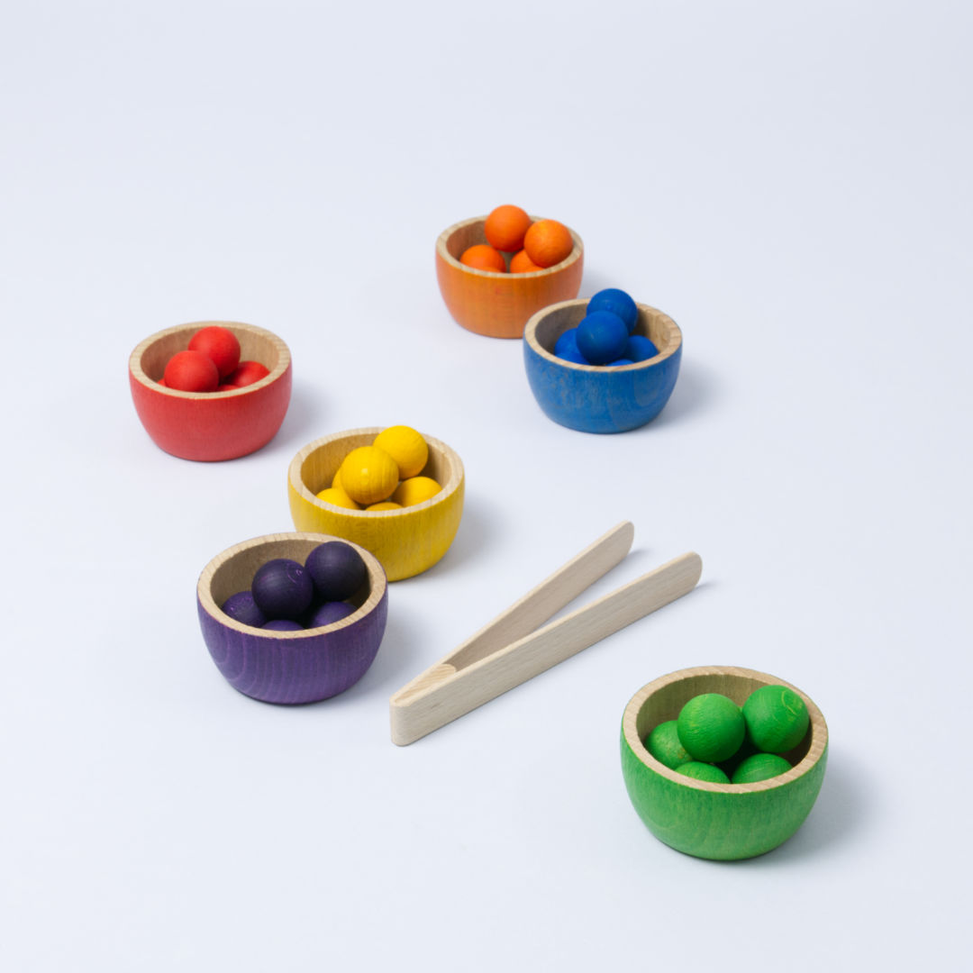 6 kleine Schüsseln in Regenbogenfarben, gefüllt mit 6 farbig passenden Kugeln, sowie eine Zange. Alles aus Holz.