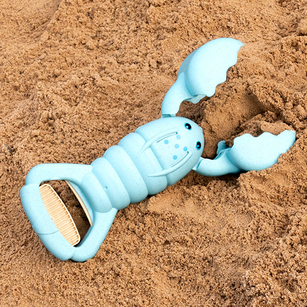 Ein hellblauer Hummer aus einem Kunststoff-Weizenstroh-Gemisch auf Sand platziert, hat einen Griff der zudrückbar ist. Die Scheren lassen somit öffnen und schließen. Er hat einen oval geformten Körper, der mit zwei Augen und fünf blauen Punkten versehen ist.