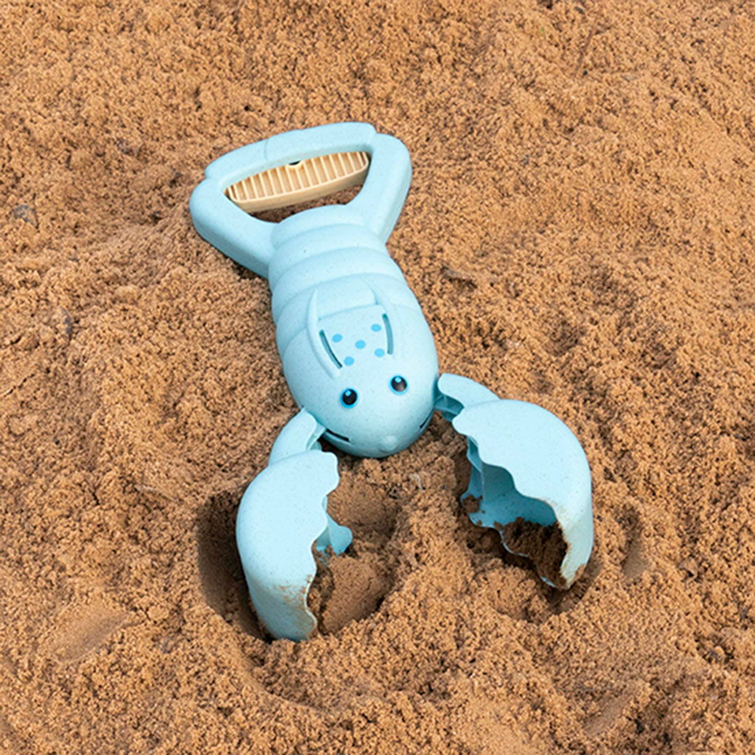 Ein hellblauer, nach vorne schauender Hummer aus einem Kunststoff-Weizenstroh-Gemisch auf Sand platziert, hat einen Griff der zudrückbar ist. Die Scheren lassen somit öffnen und schließen. Er hat einen oval geformten Körper, der mit zwei Augen und fünf blauen Punkten versehen ist.