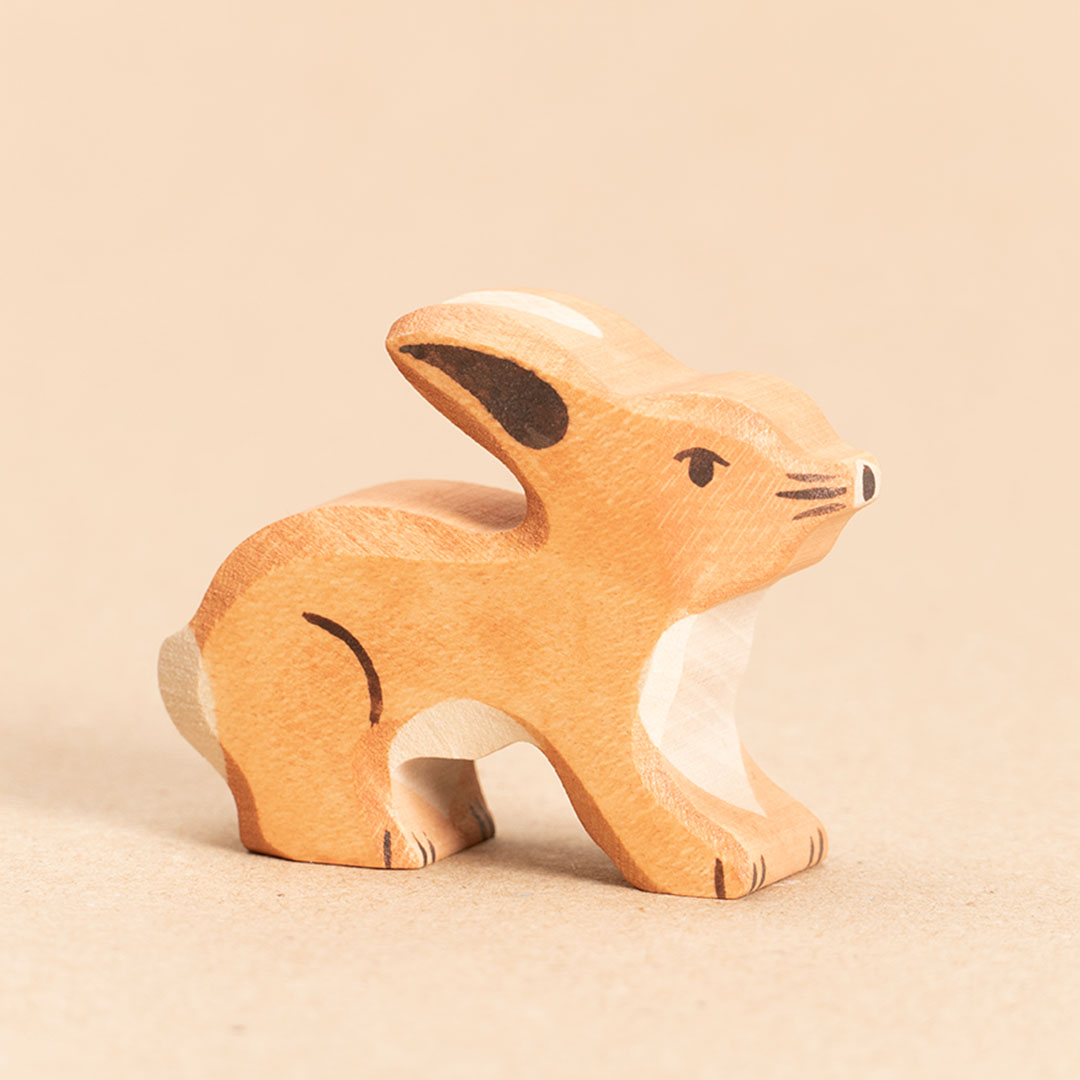 Ein stehender, hellbrauner Hase aus Ahorn- und Buchenholz gefertigt, schaut, leicht aus dem Profil nach vorne geneigt, nach rechts. Er hat ein weißes Bauchfell, sowie eine weiße Blume. Seine Ohren stehen waagerecht nach hinten.