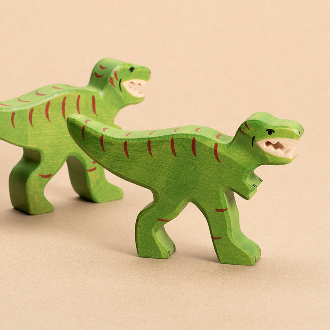 Zwei intensiv grüne Tyrannosaurus Rex aus Holz mit geöffnetem Mund und spitzen weißen Zähnen, haben rote Farbdetails, einen waagerecht nach hinten geformten Schwanz und sehr kurze nach vorne stehende Arme. Der sich im Vordergrund befindende schaut im Profil nach rechts. Der im Hintergrund schaut nach hinten rechts und ist mit seniem Schwanz im Bild angeschnitten.