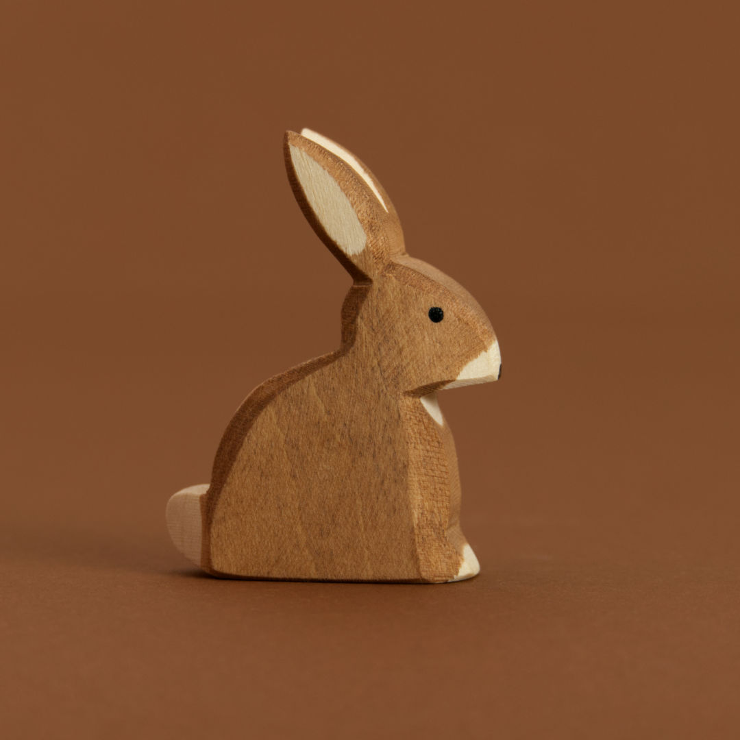 Ein brauner, sitzender Hase im Profil nach rechts schauend, ist von Ostheimer aus Naturholz gefertigt, hat eine weiße Blume und weiße Nasenspitze sowie Pfoten. Seine Ohren zeigen schräg nach oben, hinten.