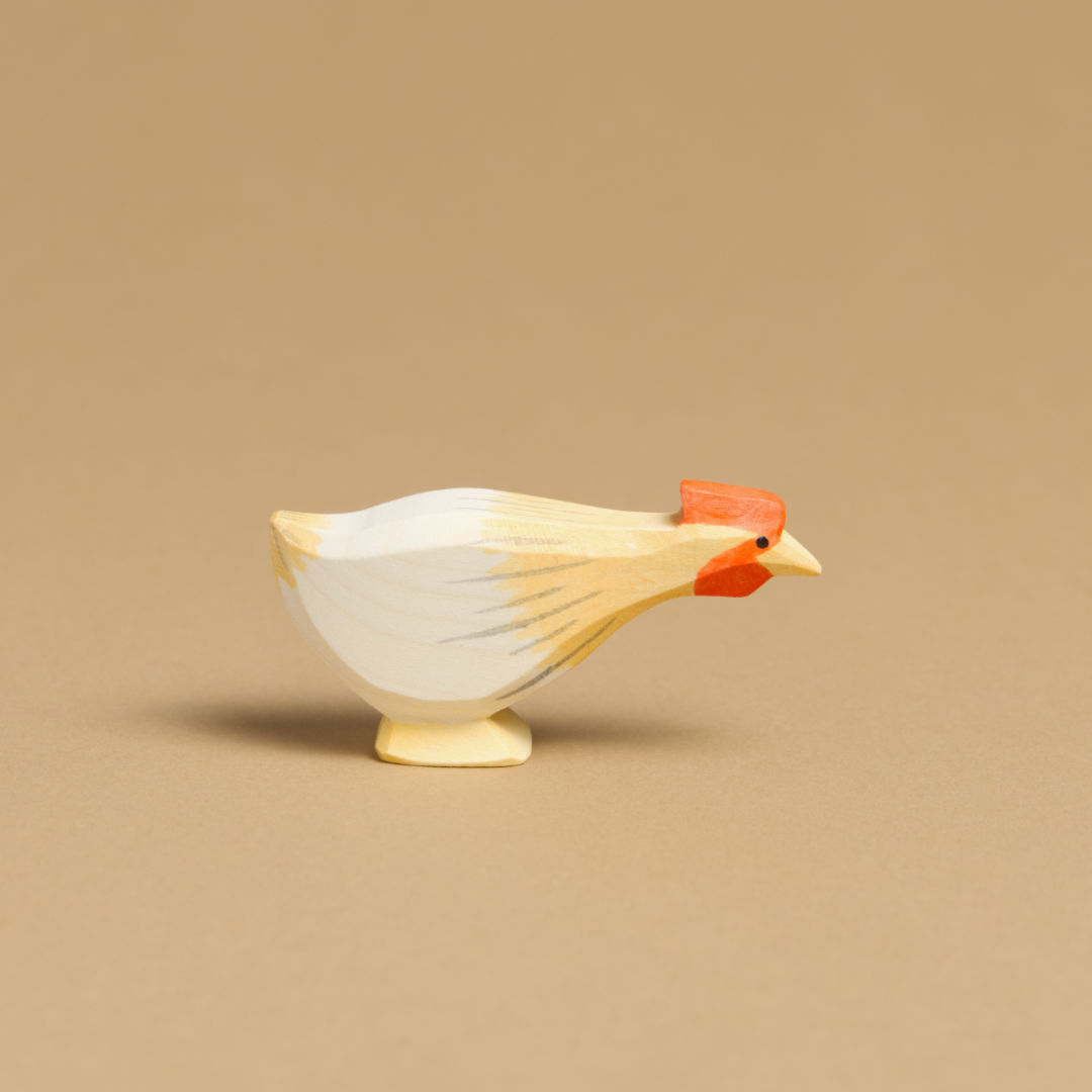 Ein langes, nach rechts im Profil schauendes Huhn von Ostheimer aus Naturholz gefertigt, ist ockerfarben, hat einen roten Kamm, weiße Federn und graue Farbdetails. Ihr Körper ist nach vorne lang gestreckt.