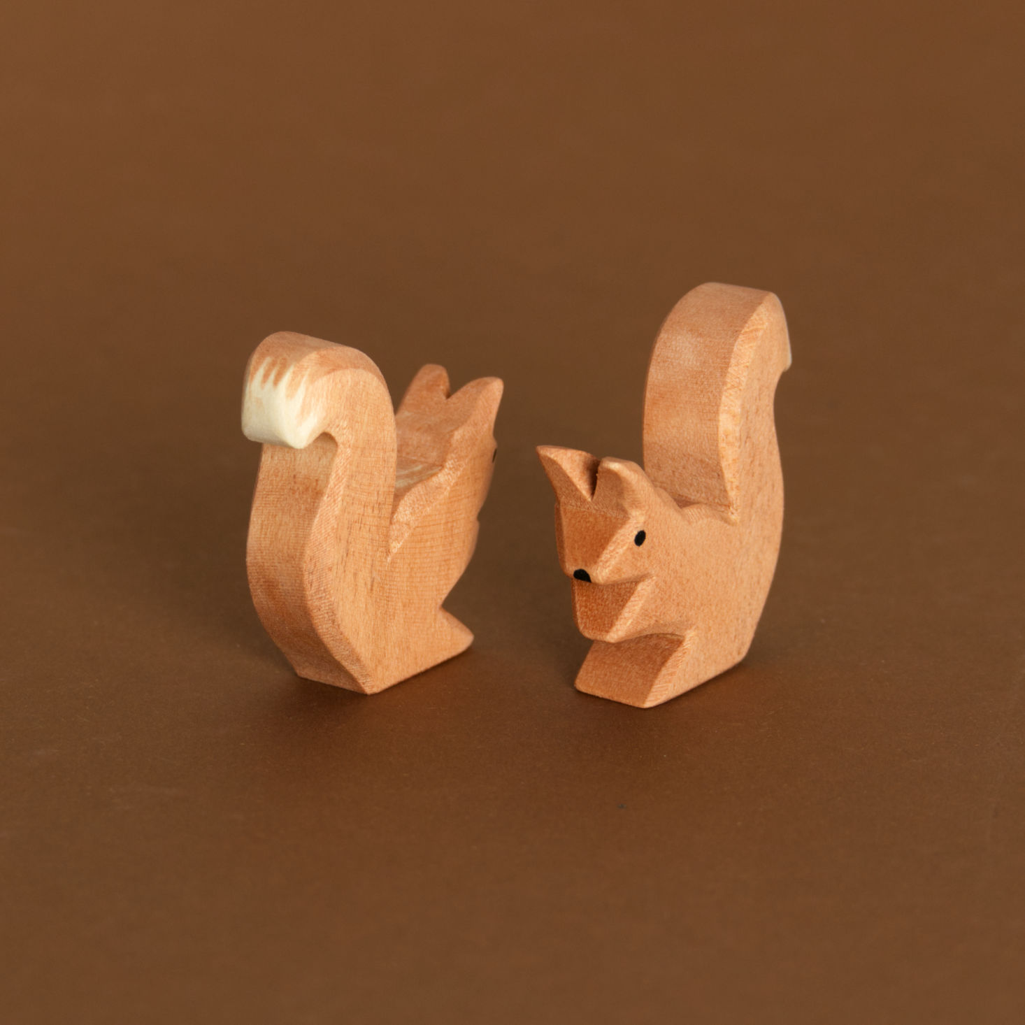 Zwei braune, sitzende Eichhörnchen aus Naturholz gefertigt von Ostheimer, sind ileicht von oben fotografiert und schauen aneinander vorbei. Das vorne rechts nach vorne links. Das links nach hinten rechts. Sie haben ihren Körper leicht nach vorne gebeugt, genauso ihre Ohren. Der Schwanz mit weißer Spitze zeigt jedoch nach oben.