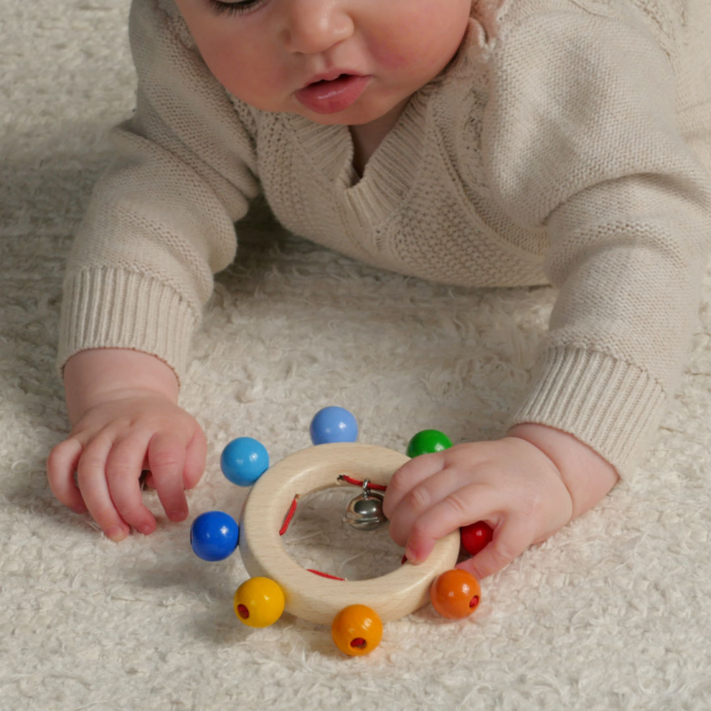 Greifling mit Regenbogenperlen, bestehend aus einem naturfarbenen Ring, der am äußeren Ringrand sich befindende bunten Perlen hat, diese immer mit gleichem Abstand angeordnet, aufweist. Im Ring Innenrand befindet sich eine in 4 Segmente eingeteilte rote Schnur. An einer dieser Segmente hängt eine metallene Glocke. Festgehalten wird der Greifling von einem Baby mit der linken Hand.