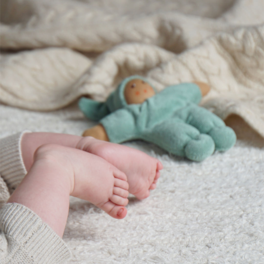 Der aus nachhaltigen Rohstoffen gefertigte Pimpel in Salbei von Nanchen mit heller Hautfarbe, ist mit Scharfswatte gestopft und liegt neben einem Kleinkind auf einem weißen Teppich und einer gestrickten Decke im Hintergrund. Im Vordergrund sind die Füße des Kindes zu sehen.
