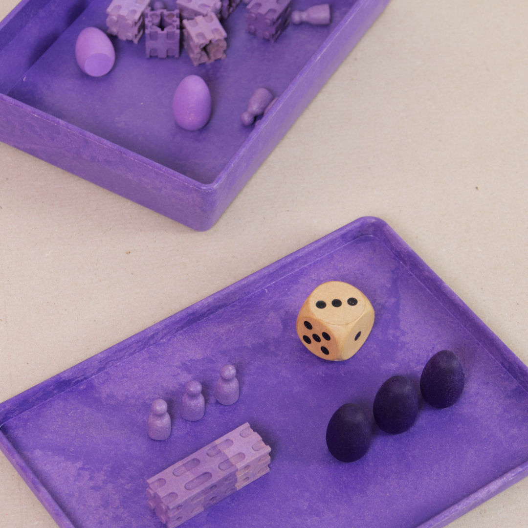 Komposition aus der aus recyclebarem Material bestehende lila Box von Wissner mit kleinen Spielsteinen und Figuren.