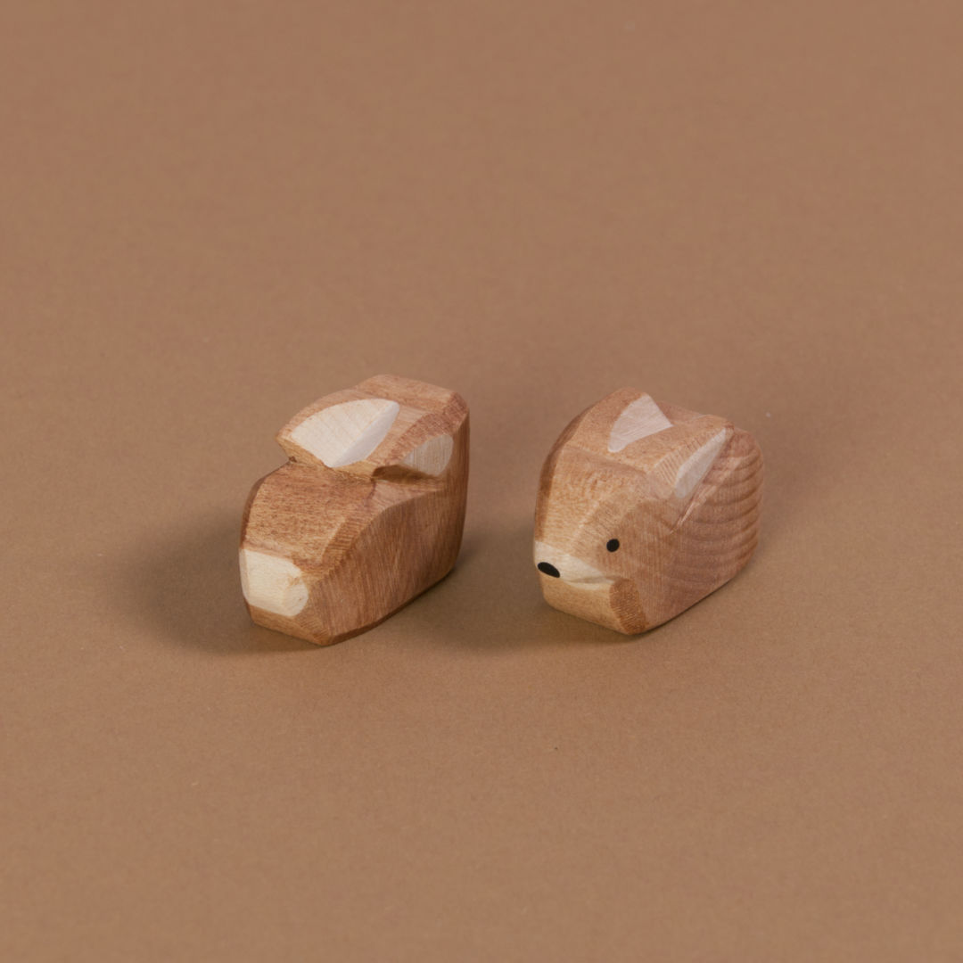Zwei aus Naturholz gefertigte Hasen von Ostheimer, schauen aneinander vorbei. Der rechte nach vorne links und der linke nach hinten rechts. Beide haben weißes Bauchfell, sowie einen weißen Schwanz. Nasenspitze und Augen sind in schwarz aufgemalt.
