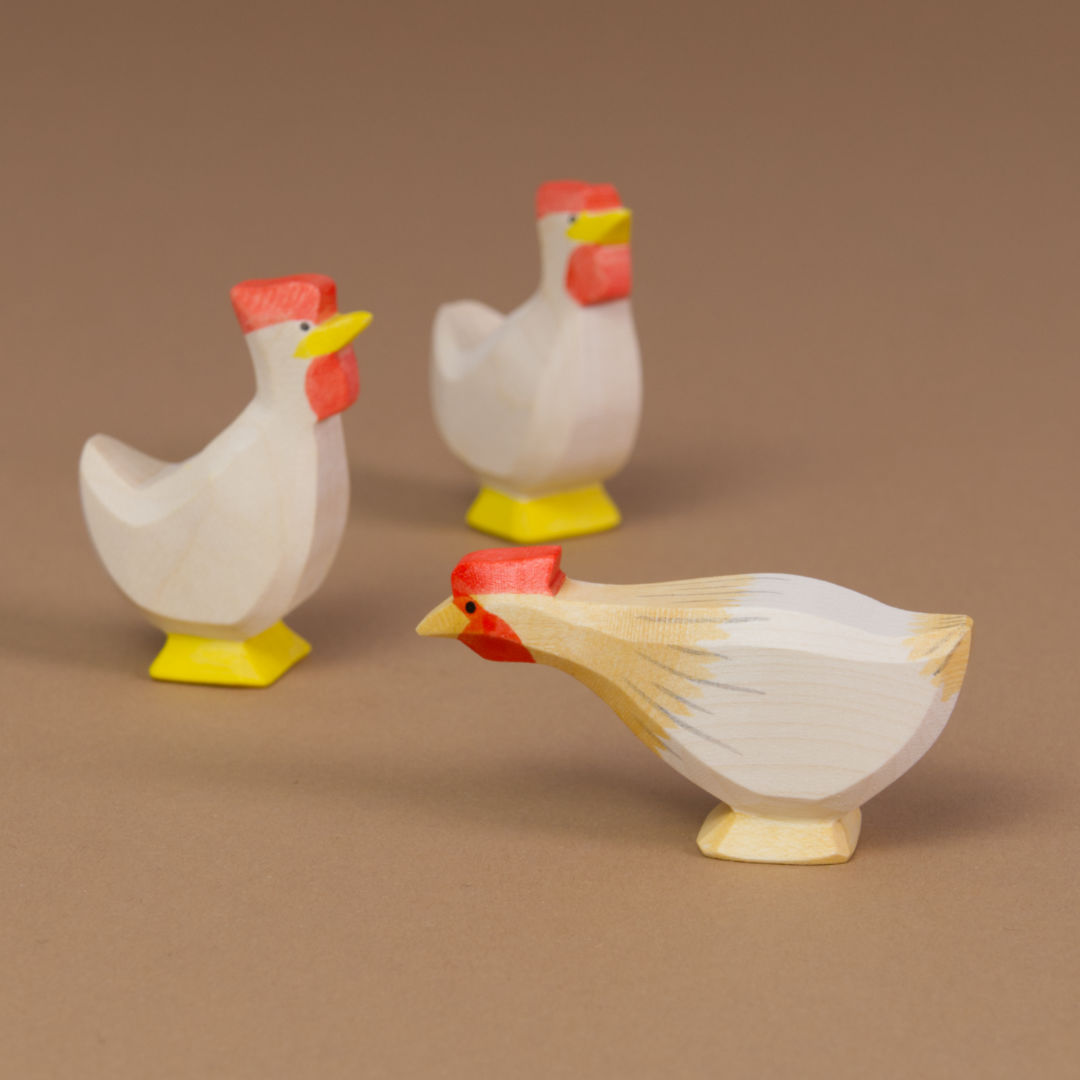 Komposition aus Huhn ocker lang und zwei Hühner in weiß. Im Vordergrund befindet sich das nach links schauende Huhn ocker lang und im Hintergrund die anderen Hühner in weiß. Alle haben einen auffällig roten Haarkamm.