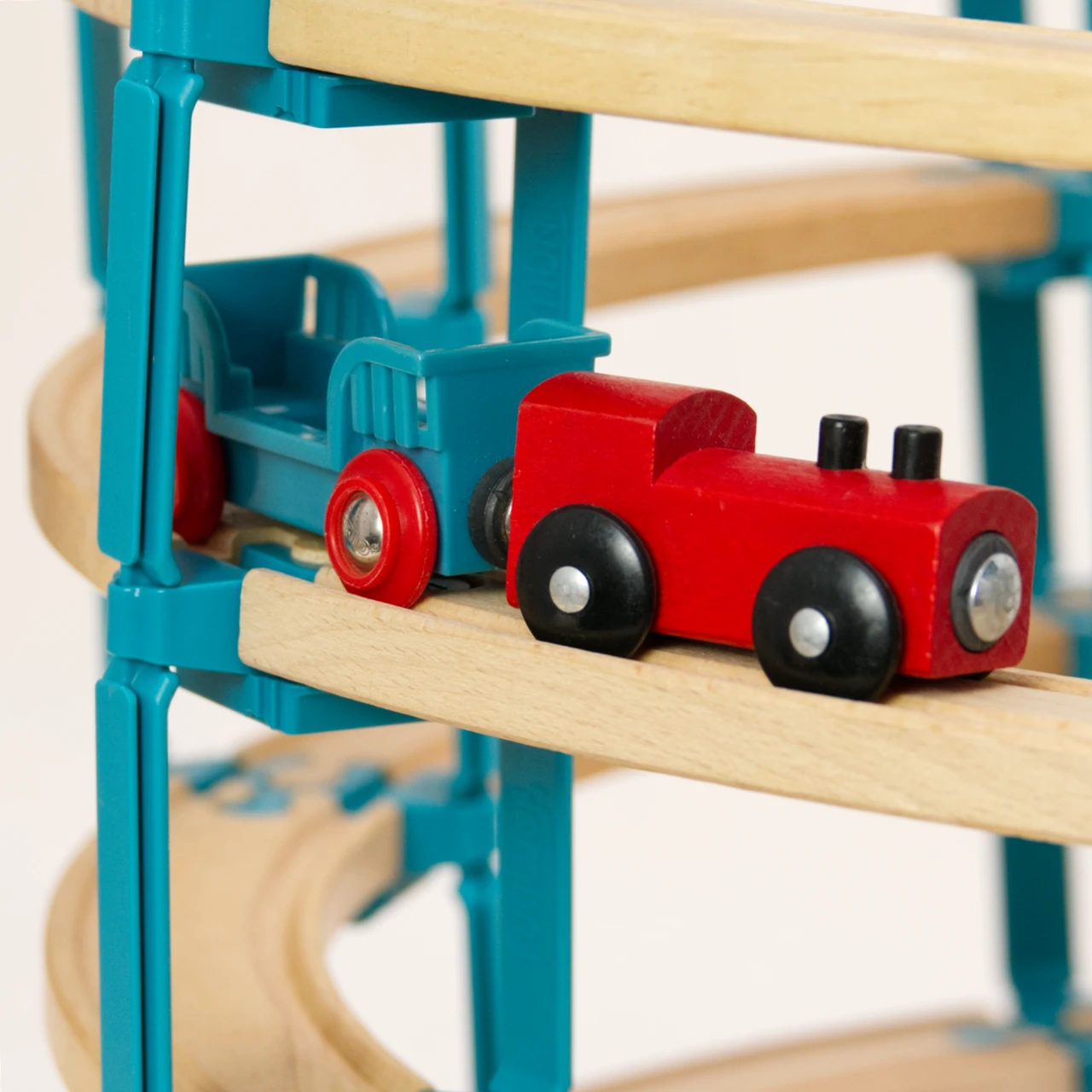 Zu sehen ist eine rote Lokomotive mit schwarzen Rädern aus Holz, an ihr hängt ein türkis farbebener Wagen. Die zwei Zugteile sind mit einem Magneten gekoppelt und stehen auf einer mehrstöckigen Spielzeug Holzschienebahn. Die Schienen sind mit den Verbindungselementen LinkUp und Lift aus dem Helix Set von Yamba zusammengebaut.