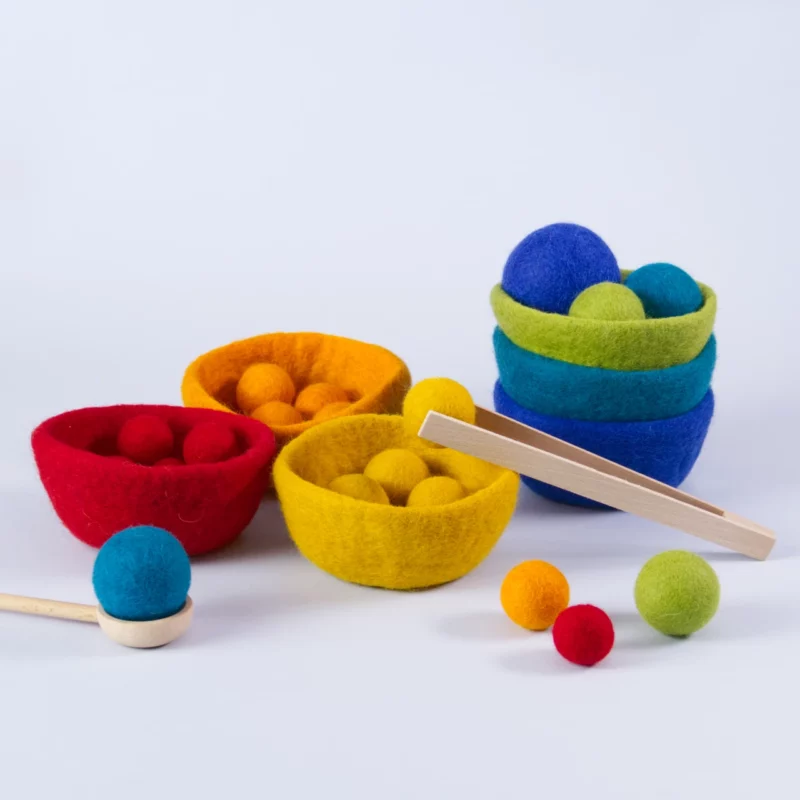 Sortierspiel Filz sorterino goki evolution mit sechs farbigen Filzschalen, farblich passenden Filzkugeln in Regenbogenfarben, Holzzange und Holzlöffel – nachhaltiges Montessori-Spielzeug zur Förderung von Feinmotorik und Farbzuordnung. Hier im Aufbau sind einige Schalen in einander gesteckt, ein Teil der Kugeln ist einsortiert. Die Holzpinzette hält einen der gelben Bälle, auf dem Löffel liegt ein Bällchen in der Farbe Türkis.