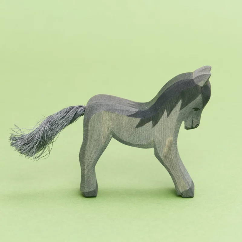 Ein hölzernes Fohlen der Marke Ostheimer, das nach rechts schaut. Die flach ausgesägte Figur zeigt die Silhouette eines stehenden Fohlens, die Kanten sind leicht abgerundet. Das Fell ist dunkelgrau und die Mähne ist schwarz. Der Schweif ist eine graue Kordel.