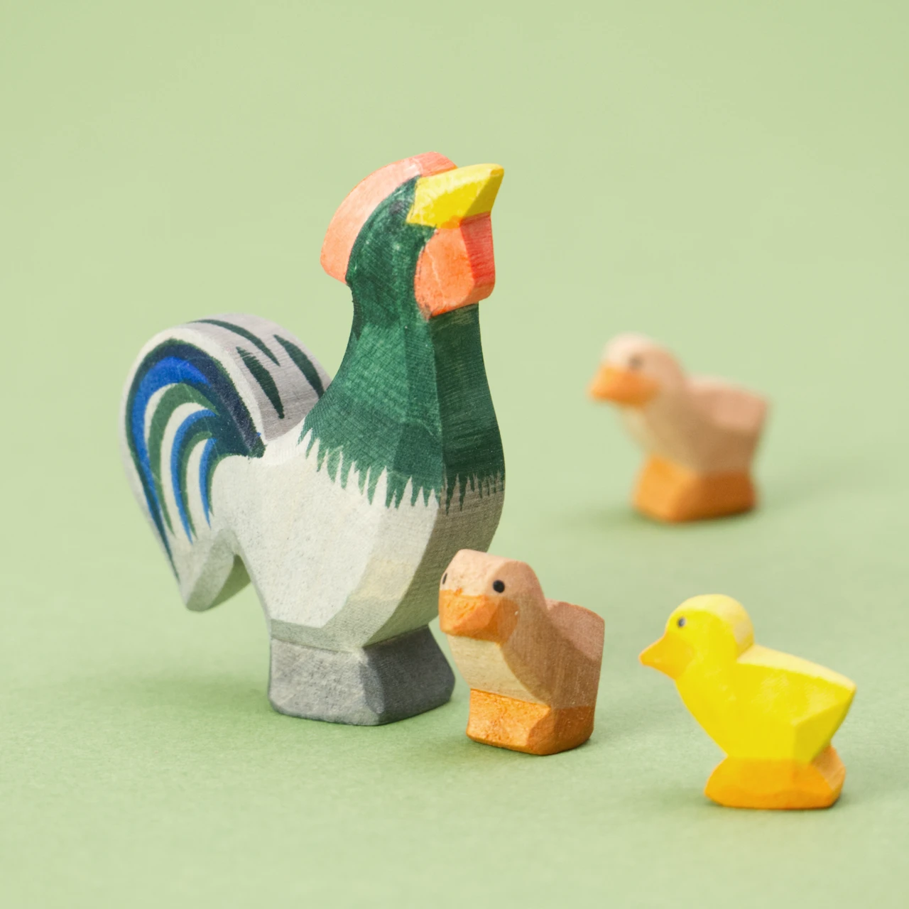 Ein hölzerner Hahn der Marke Ostheimer mit drei hölzernen kleinen Kücken, eins ist gelb, die anderen beiden sind braun. Der Hahn hat einen grau grünes Gefieder mit blauen Schwanzfedern. Er hat einen gelben Schnabel und einen roten Kamm, die Augen bestehen aus kleinen schwarzen Punkten.