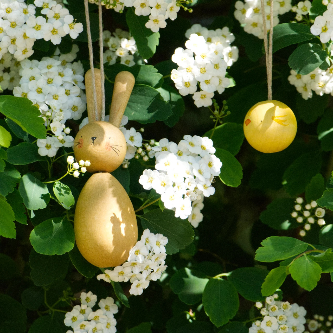 Hop and Peep von Grapat aus Holz, besteht aus einem braunen Hasen und gelben Küken zum Aufhängen, hier hängen diese in einem Busch mit Blumen.