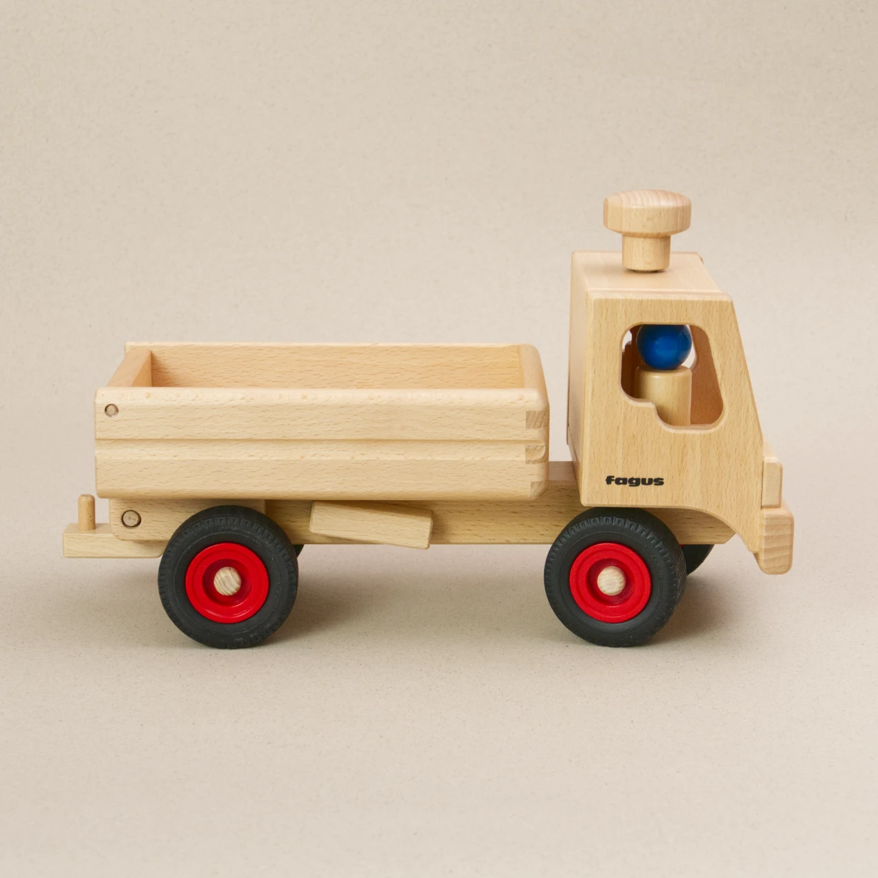Holzfahrzeug LKW Kipper von fagus aus naturlackiertem Buchenholz mit schwarzen Vollgummireifen und roten Radkappen sowie großer Ladefläche. Seitenansicht, in der Fahrerkabine sitzen zwei Figuren, auf dem Dach ist ein Lenkknopf.